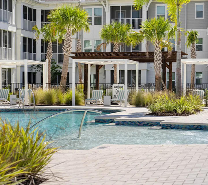 Luxurious apartment complex featuring a sparkling pool surrounded by palm trees, elegant cabanas, and lush greenery.