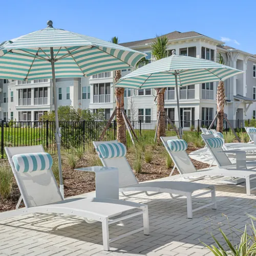 Luxurious apartment complex with pristine poolside lounging area, featuring stylish sun chairs and striped umbrellas under a clear blue sky.