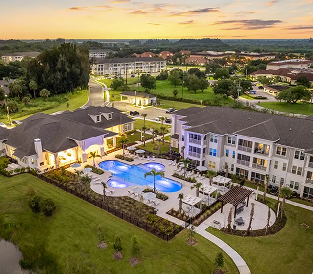 Aerial view of a luxurious apartment complex with beautifully landscaped gardens, a sparkling pool, and modern amenities at sunset.