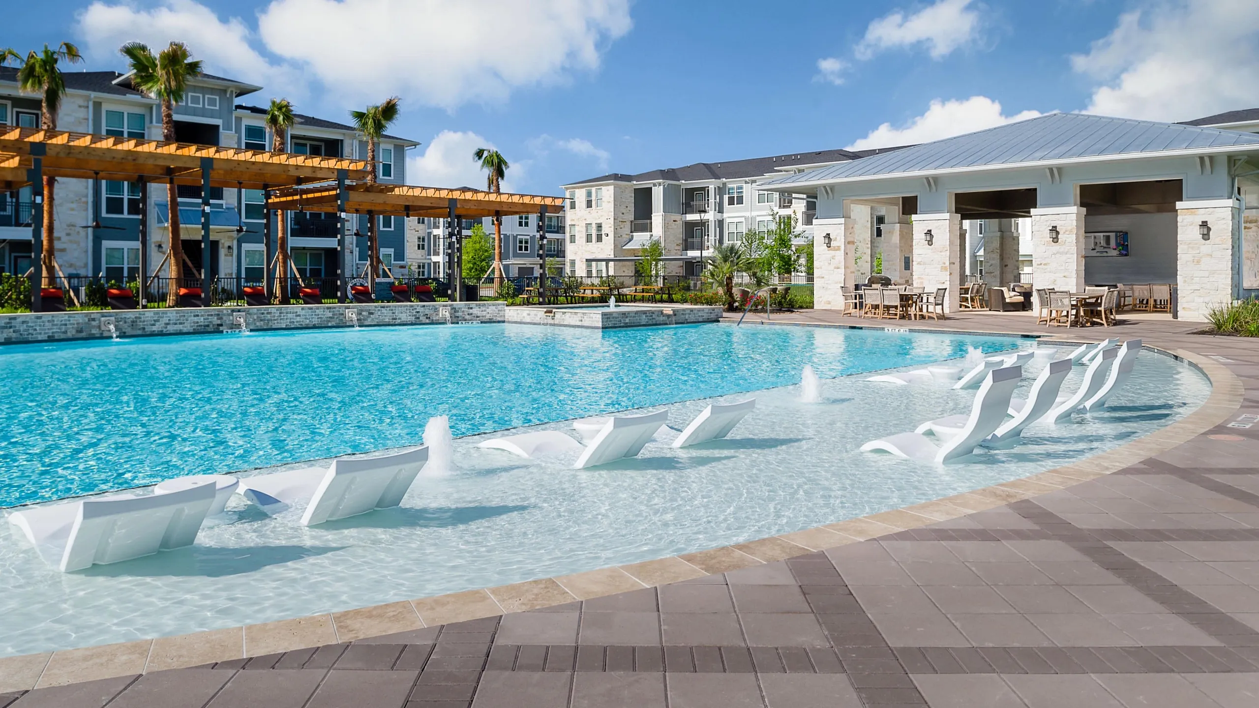 Luxurious apartment complex featuring a stunning outdoor pool with stylish lounge chairs, palm trees, and a beautifully designed patio area.