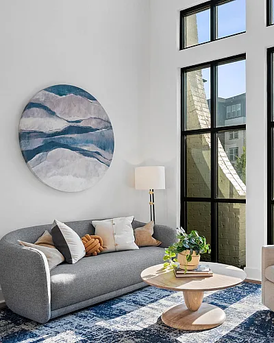 Elegant living room with high ceilings, featuring a modern gray sofa, plush armchair, and a stylish round coffee table by large, sunlit windows.