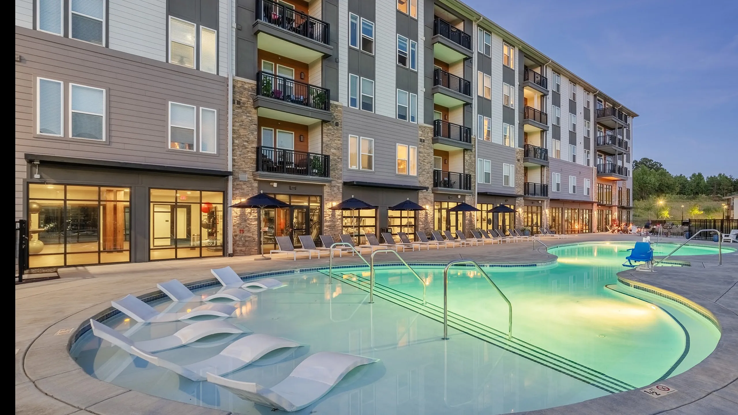 Elegant apartment complex featuring a beautifully illuminated pool area with lounge chairs and modern architecture.
