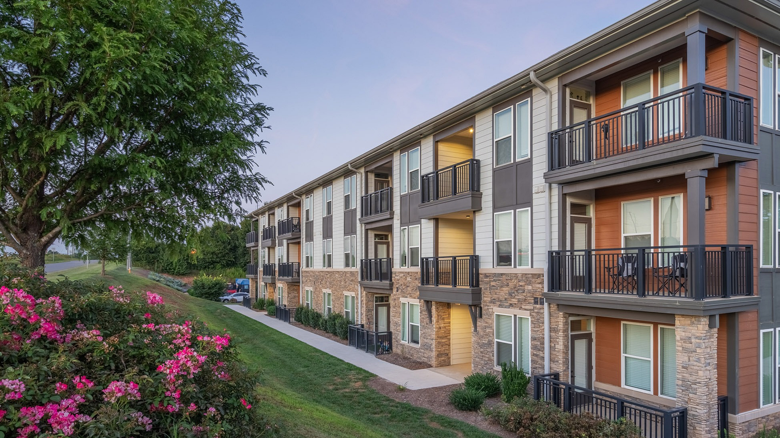 Charming three-story apartment building with modern balconies, surrounded by lush greenery and vibrant pink flowers.
