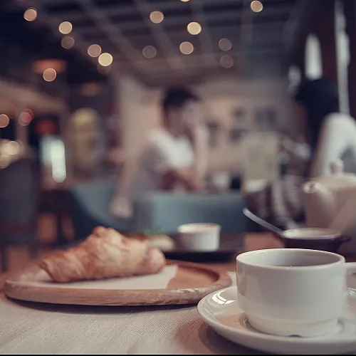 Cozy caf&eacute; atmosphere with a close-up of a steaming cup of coffee and a freshly baked croissant on a wooden tray. Soft bokeh lights in the background create a warm and inviting ambiance.
