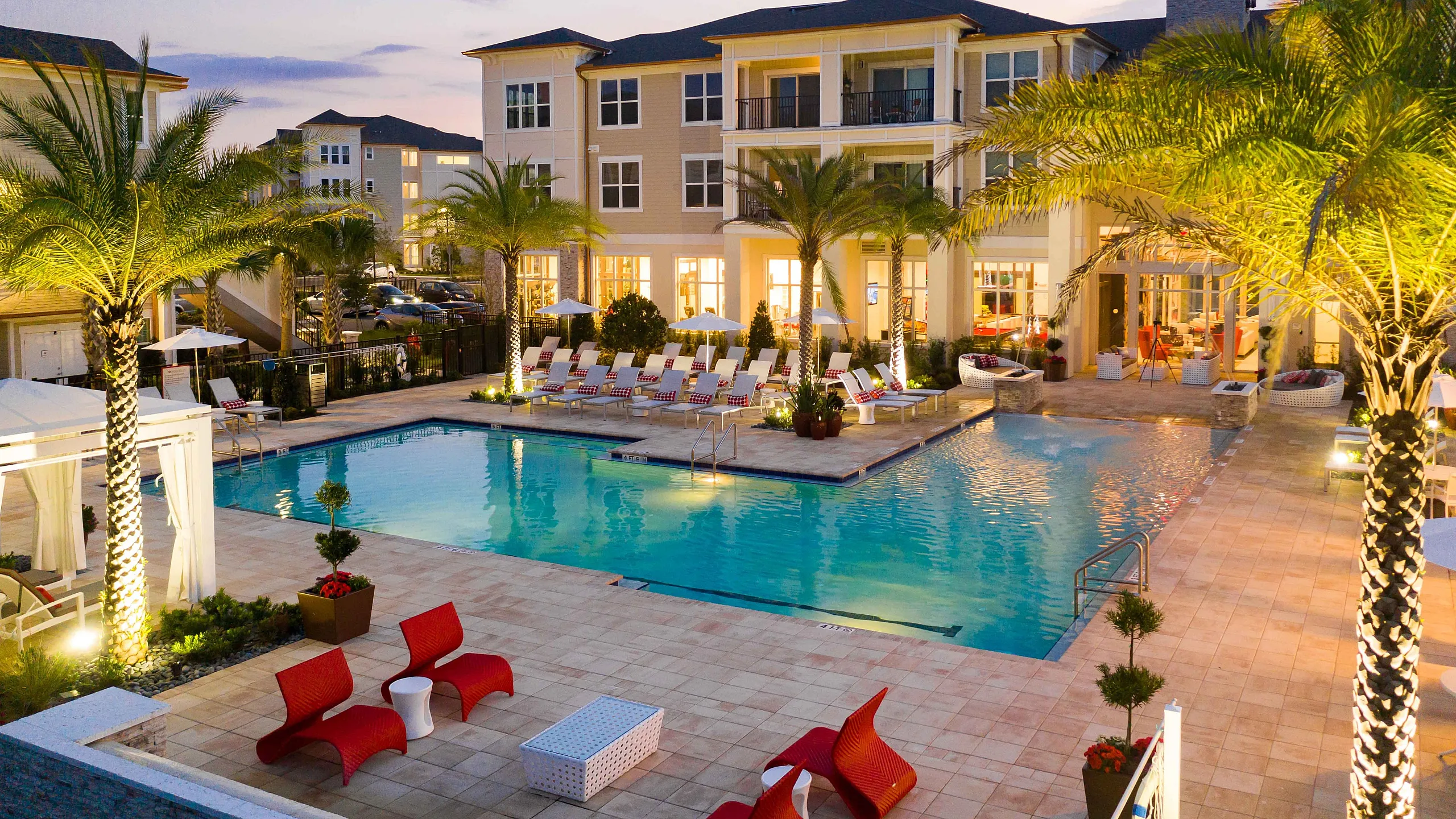 Luxurious apartment complex with a sparkling pool, lush palm trees, and modern seating under a twilight sky.