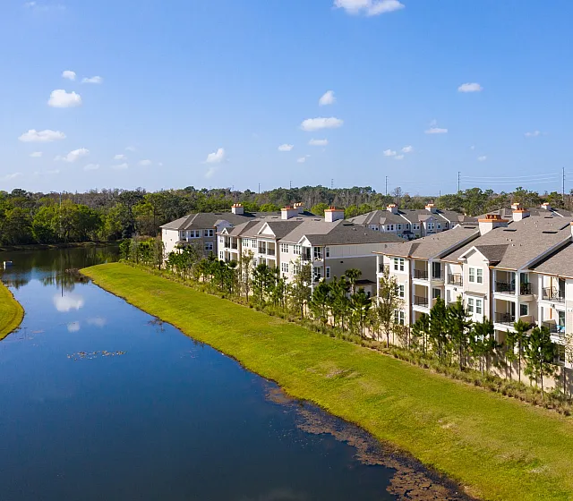 Charming lakeside apartment complex with lush landscaping and serene water views under a bright blue sky.