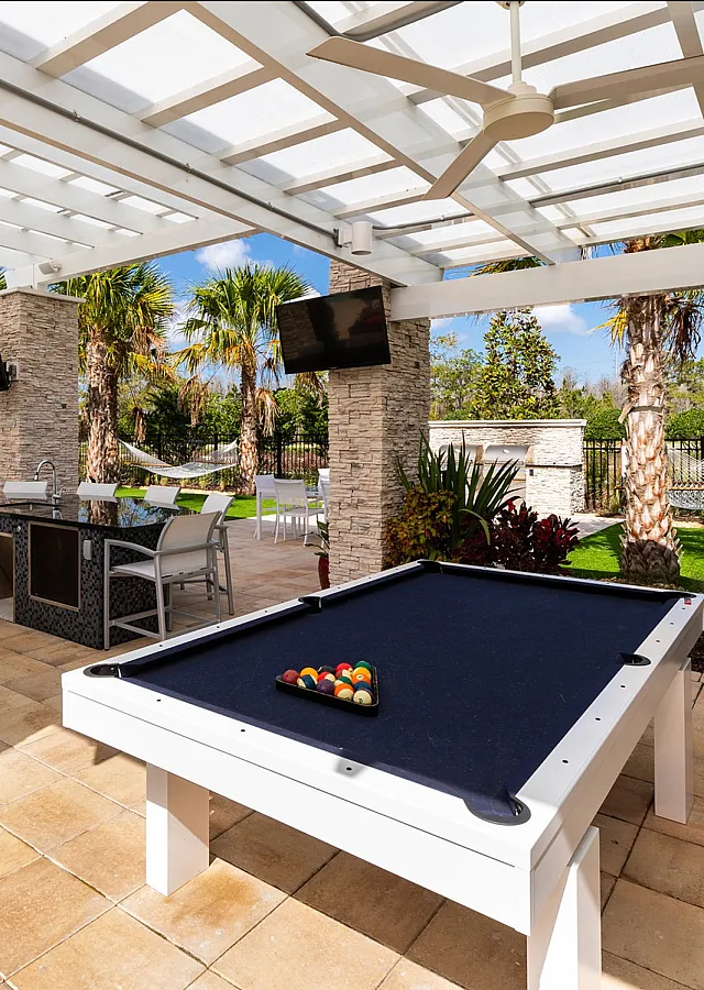 Luxurious outdoor recreational area featuring a modern pool table, shaded patio with sleek seating, multiple flat-screen TVs, and a lush garden backdrop.