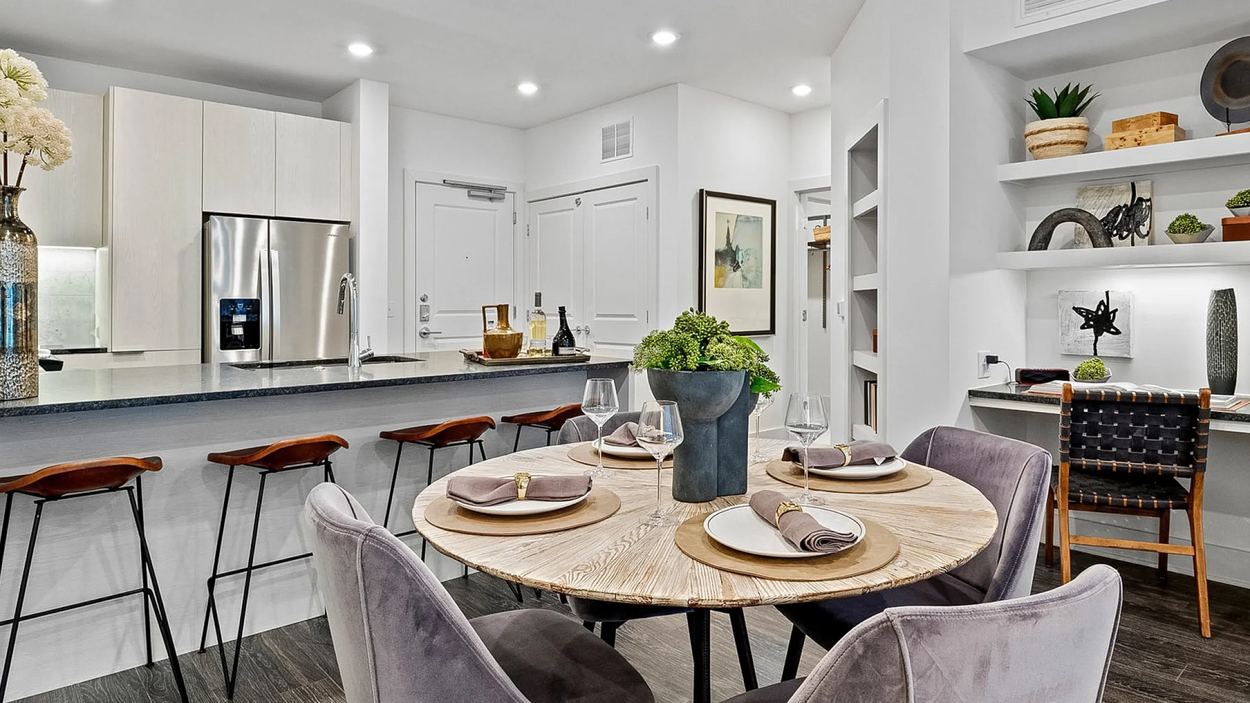 Elegant open-concept dining and kitchen area with modern decor, featuring a round wooden table, plush grey chairs, and a sleek breakfast bar with stylish stools.
