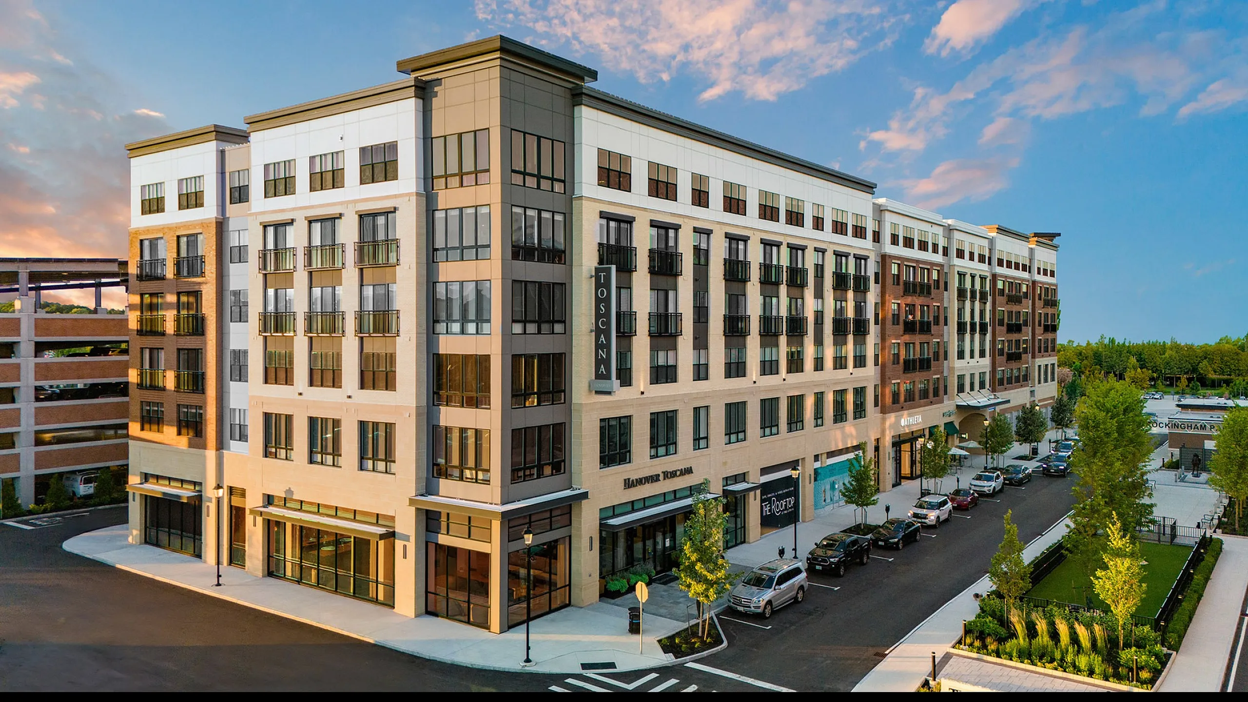 Elegant exterior view of a modern apartment complex with large windows, lush greenery, and ample parking, set against a vibrant evening sky.