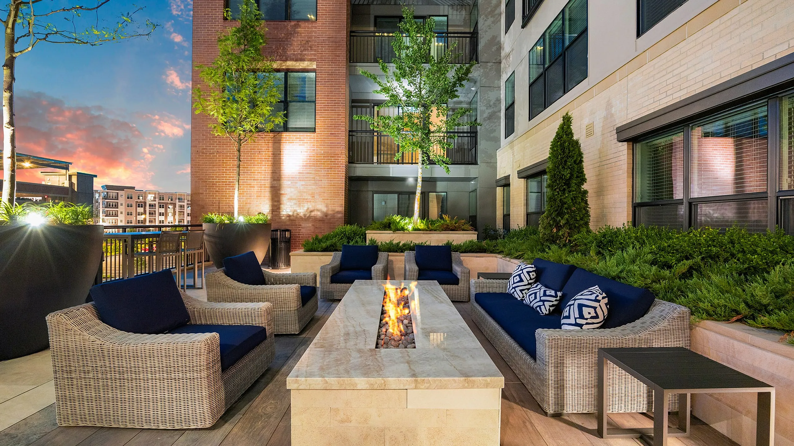 Luxurious outdoor patio with elegant wicker furniture, plush navy cushions, and a modern fire pit, offering a serene ambiance under a twilight sky.