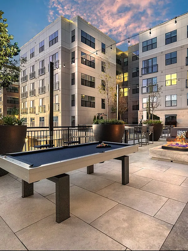 Elegant urban courtyard at dusk featuring a modern pool table, cozy fire pit, and chic seating area with cushions, surrounded by lush greenery and a stylish apartment complex.