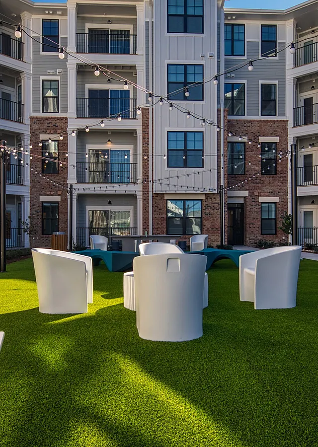 Charming apartment courtyard with lush green lawn, modern seating, and elegant string lighting against stylish brick and white facade.