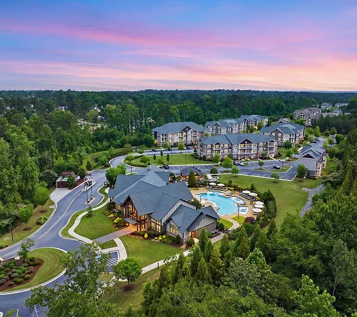 Luxurious apartment complex featuring beautifully landscaped grounds, a resort-style pool, and modern architecture surrounded by lush greenery at sunset.