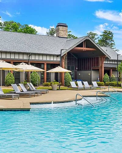 Luxurious apartment community featuring a sparkling pool, elegant lounge chairs, and lush landscaping under a vibrant blue sky.