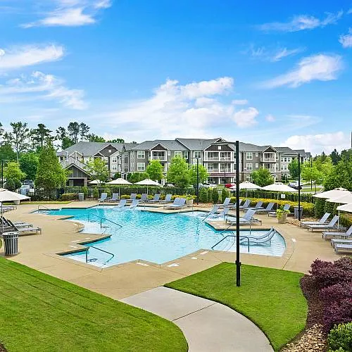 Luxurious apartment complex featuring a sparkling pool surrounded by elegant lounge chairs and lush landscaping, under a bright blue sky.