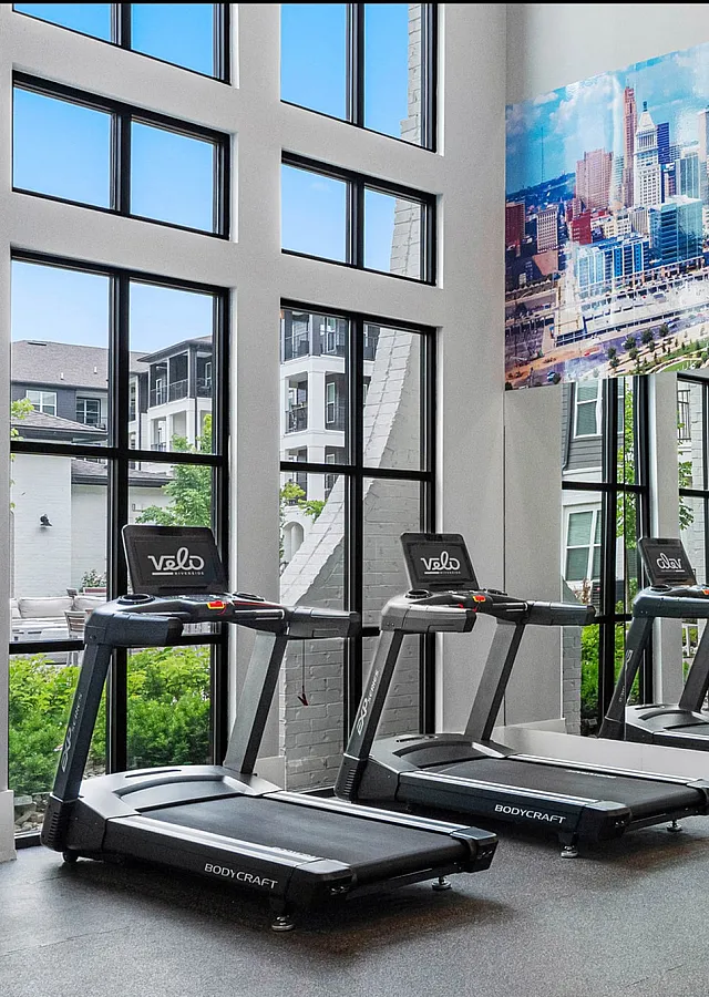 Modern fitness center with floor-to-ceiling windows, offering treadmills and a view of the city skyline mural.