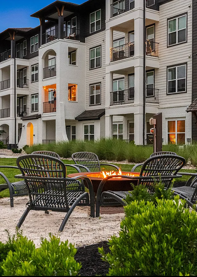 Modern apartment complex with cozy outdoor fire pit area, surrounded by lush greenery and seating, perfect for social gatherings.
