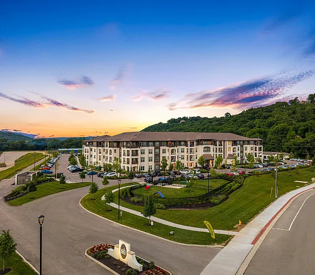 Stunning aerial view of a modern apartment complex nestled in lush greenery with a picturesque sunset sky, highlighting serene living and convenient location.