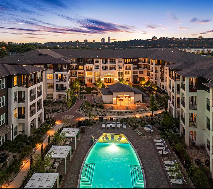 Luxurious apartment complex with a stunning illuminated pool at dusk, surrounded by lush landscape and modern architecture.
