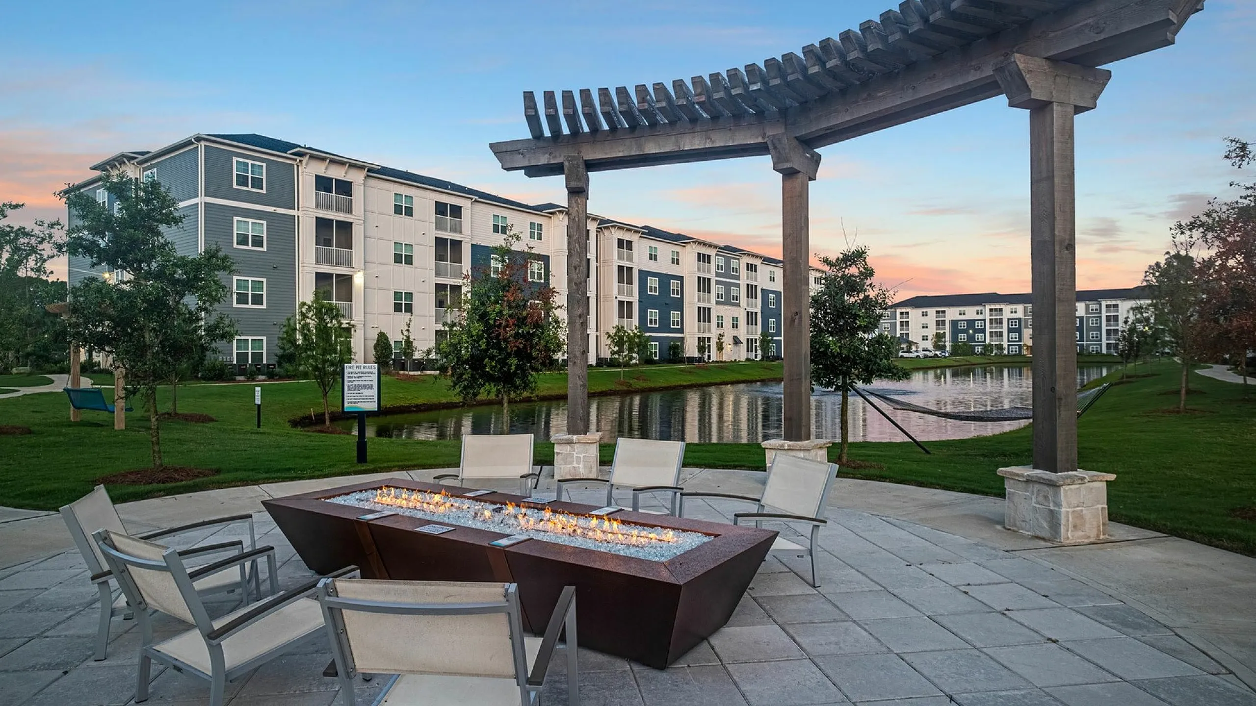 Charming apartment complex featuring modern outdoor seating with a cozy fire pit, surrounded by lush greenery and a serene pond at sunset.