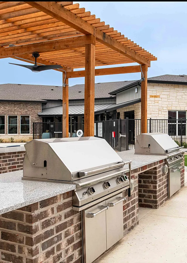Modern outdoor kitchen with sleek stainless steel grills under a charming wooden pergola, adjacent to a vibrant community clubhouse.