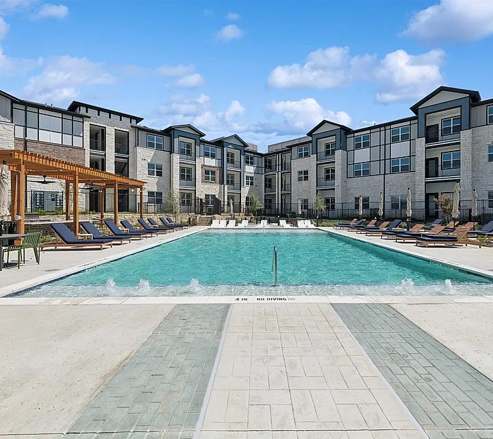 Luxurious apartment complex with a glistening pool, stylish sun loungers, and modern architecture under a bright blue sky.