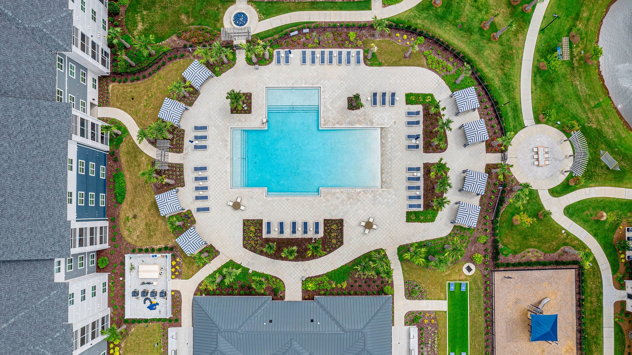 Aerial view of luxurious apartment complex showcasing a stunning central pool with sun loungers, surrounded by vibrant green landscapes and modern amenities, offering a perfect blend of relaxation and upscale living.