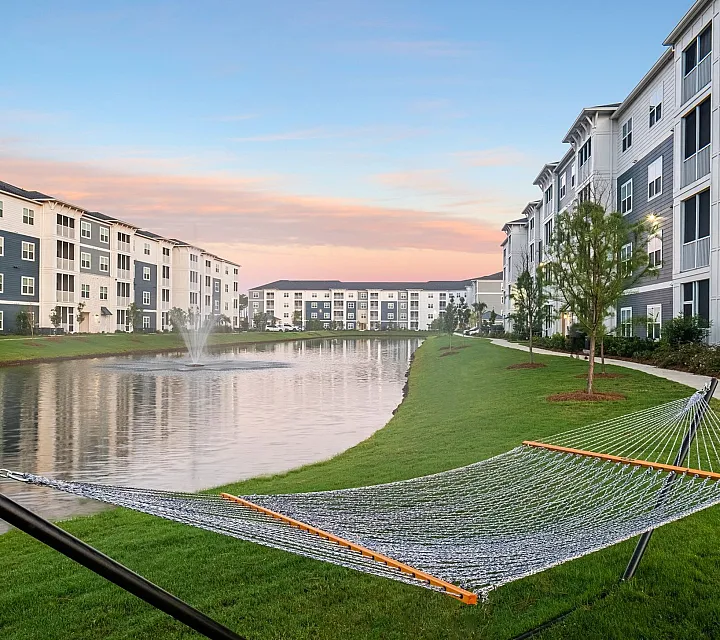 Charming apartment complex surrounded by lush greenery and a serene lake with a fountain, offering a tranquil lifestyle.