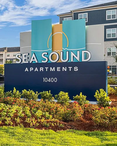 Inviting entrance to Sea Sound Apartments with vibrant landscaping and modern architecture under a clear blue sky.