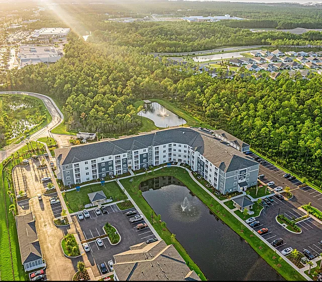 Stunning aerial view of a modern apartment complex nestled beside lush greenery, featuring a serene pond with fountains and ample parking space, basking in warm sunlight.
