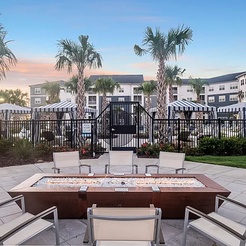 Luxurious apartment complex pool area at sunset, featuring elegant palm trees, stylish cabanas with striped awnings, and comfortable seating around a sleek fire pit.
