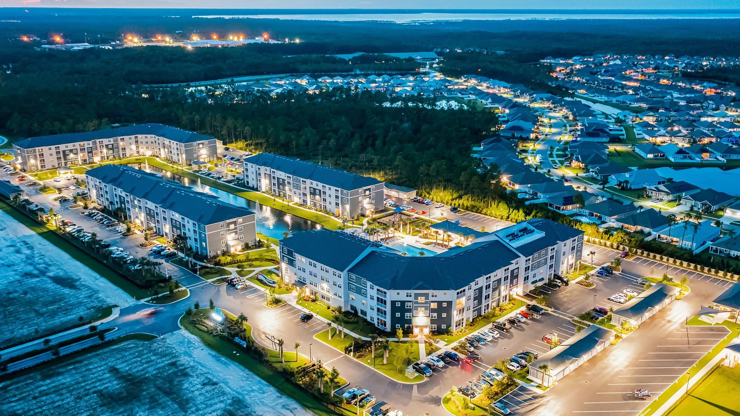 Aerial view of a vibrant apartment complex at dusk, showcasing modern architecture, beautifully landscaped grounds, and ample parking.