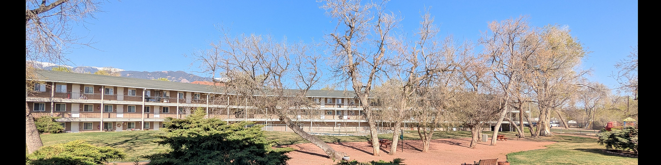 Charming apartment complex with a lush courtyard, vibrant greenery, and picturesque trees under a clear blue sky.
