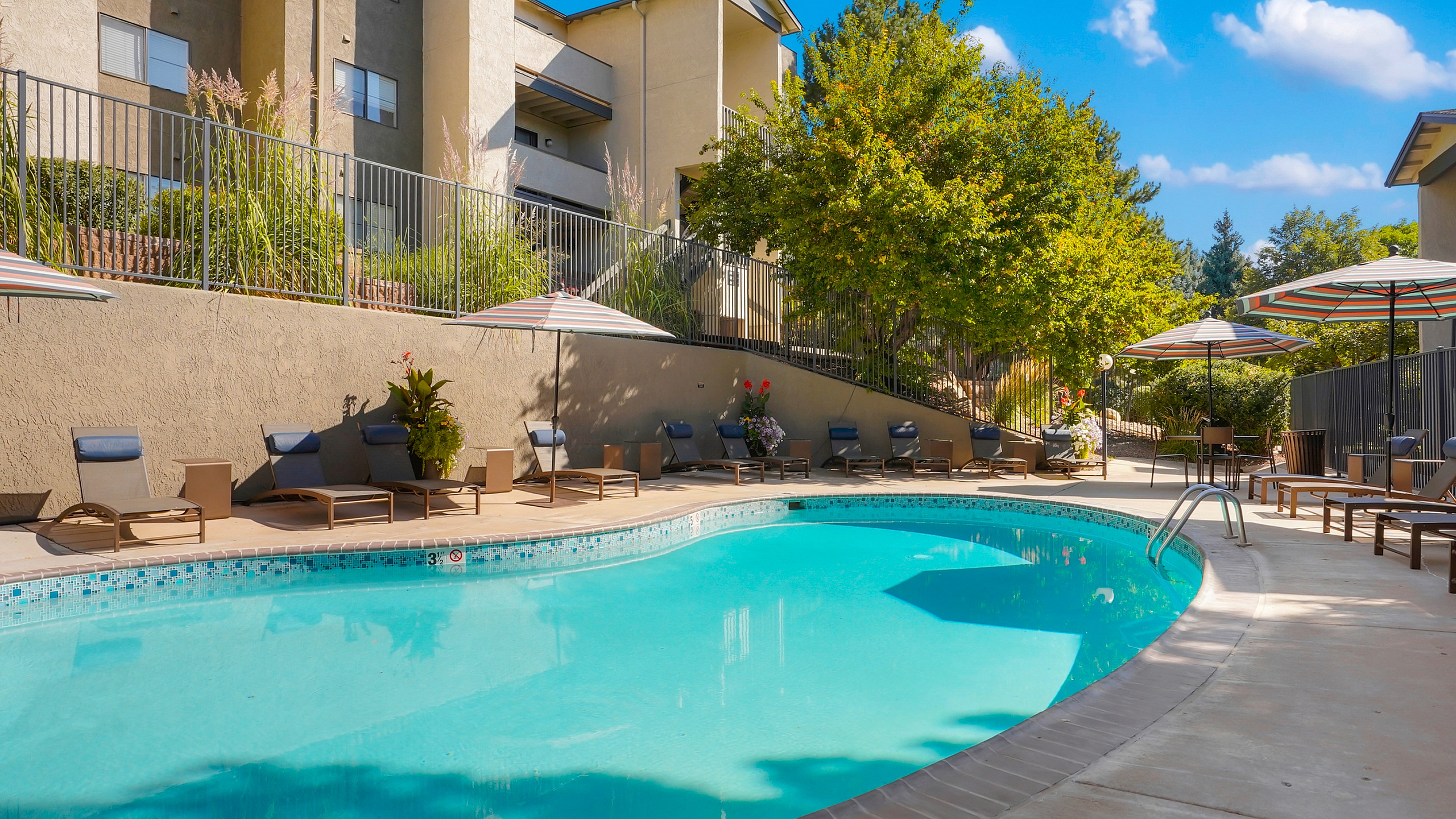 Inviting outdoor pool area with modern lounge chairs and striped umbrellas, surrounded by lush greenery and stylish architecture under a clear blue sky.