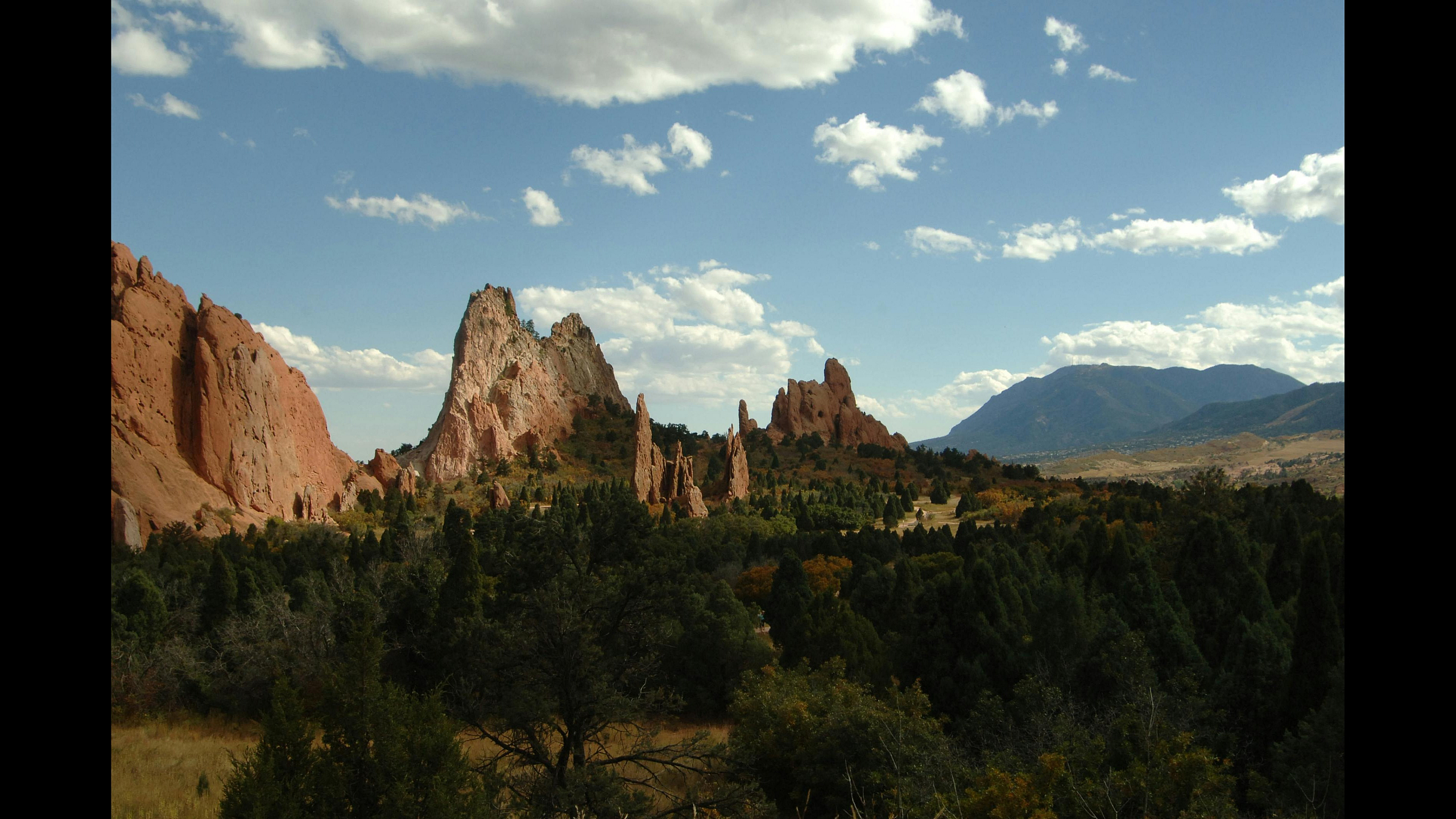 Breathtaking landscape with towering red rock formations under a blue sky, surrounded by lush greenery and distant mountains.