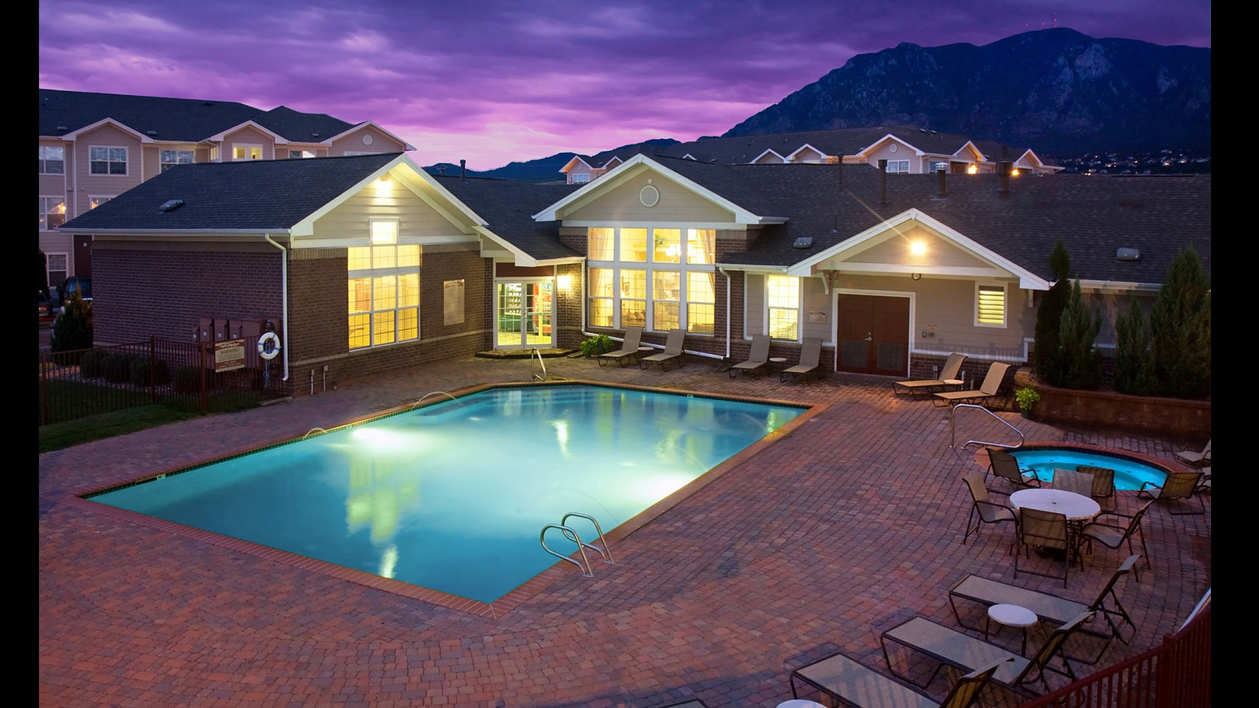 Charming mountain-side apartment complex featuring a beautifully illuminated pool and spa area at dusk, surrounded by inviting lounge chairs and scenic views.