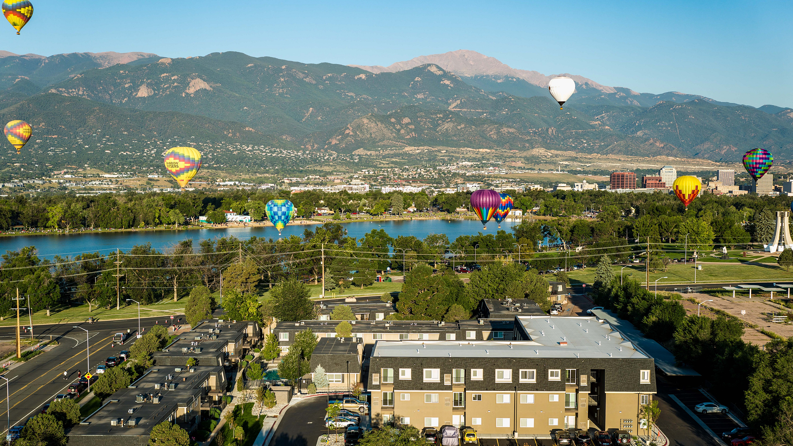 Stunning aerial view of a vibrant neighborhood with scenic mountain backdrop and colorful hot air balloons soaring above a serene lake.