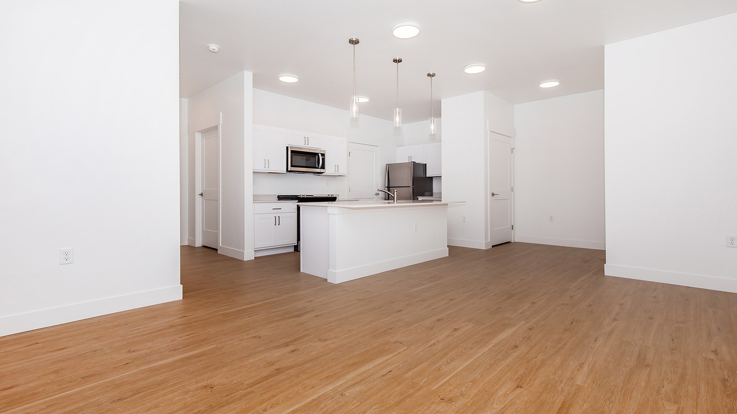 Modern open-concept kitchen with sleek white cabinets, stainless steel appliances, and elegant pendant lighting, set against warm wood flooring.