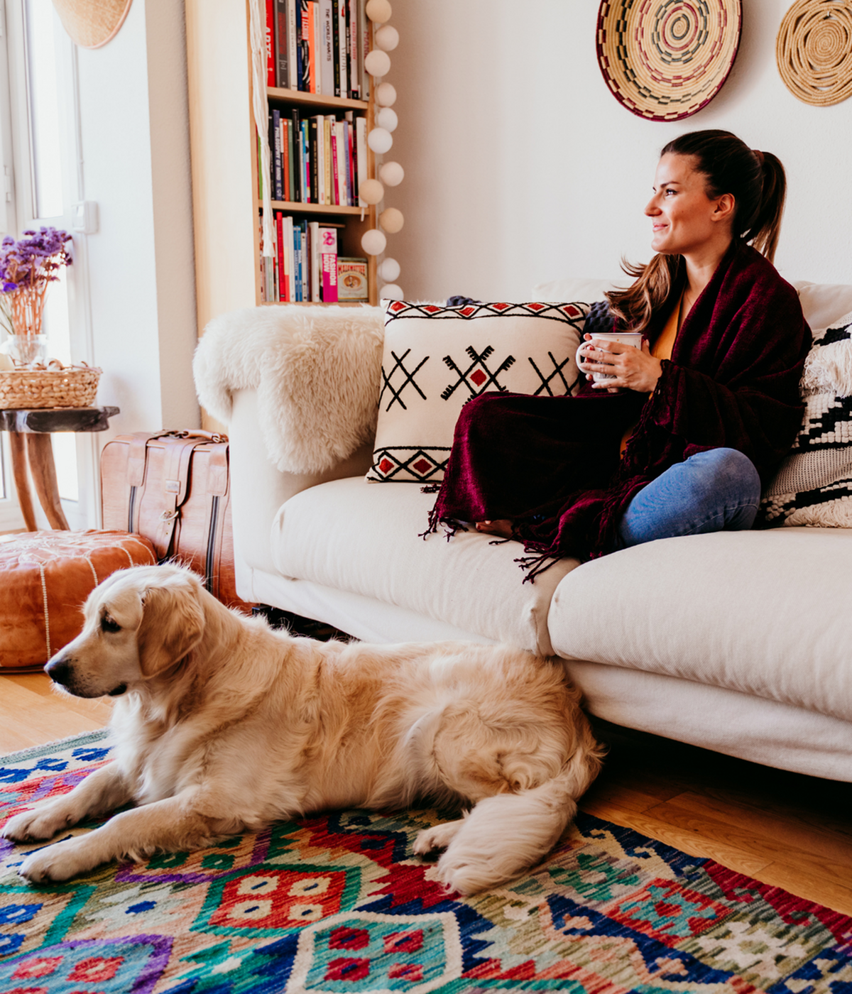 Cozy living room with vibrant rug, a comfortable couch, and a golden retriever; a woman enjoying a warm drink, surrounded by stylish decor and a bookshelf.