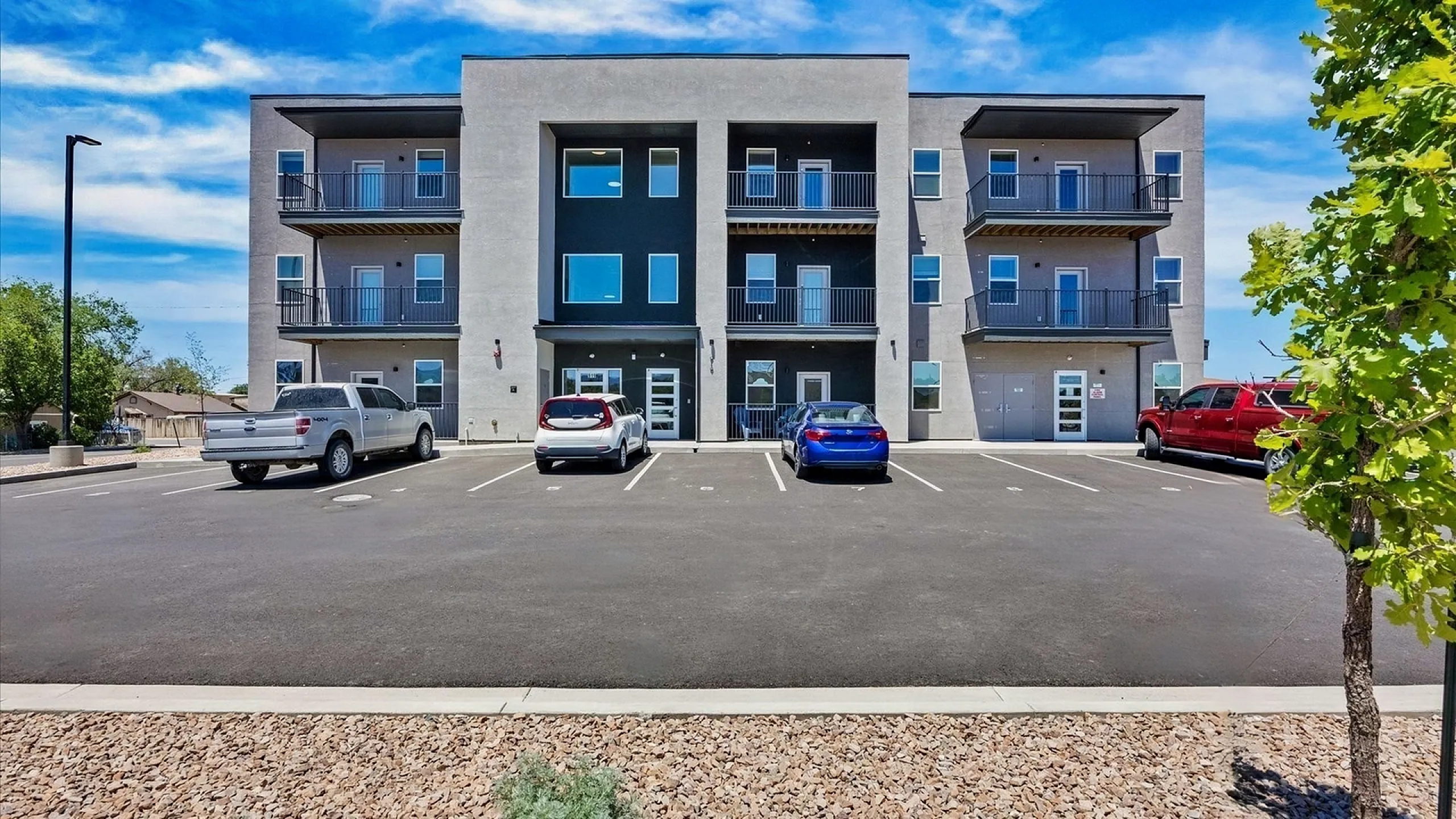 Modern three-story apartment building with spacious balconies and ample parking, set against a clear blue sky.