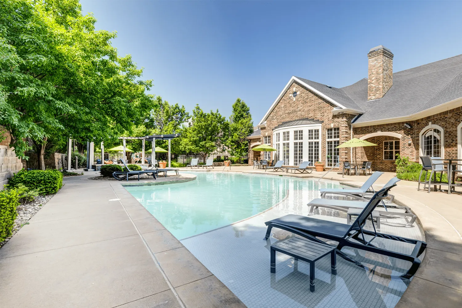 Luxurious outdoor pool area with stylish loungers, surrounded by lush greenery and a charming brick clubhouse.