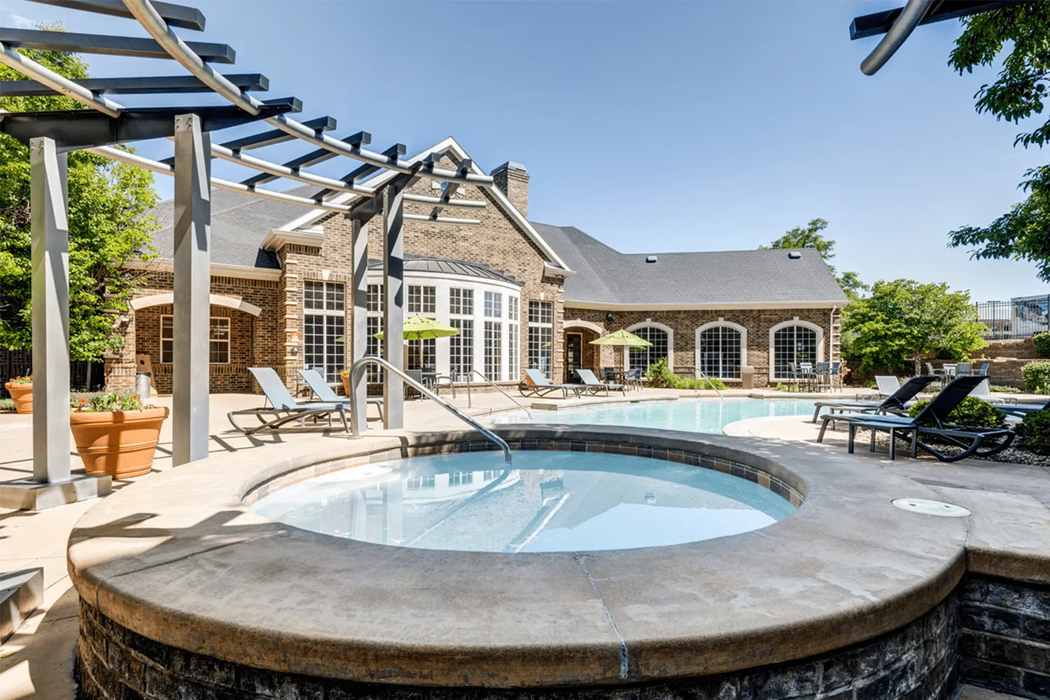 Luxurious outdoor pool and spa area featuring elegant lounge chairs and lush greenery, perfect for relaxation by the brick clubhouse.