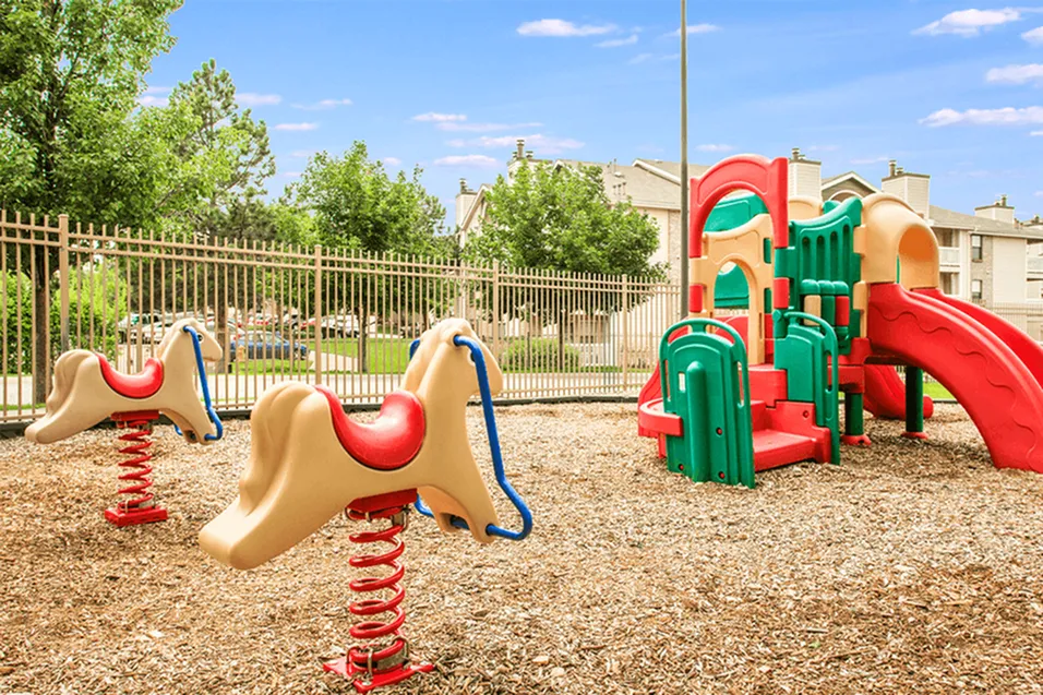 Inviting children\'s playground with colorful slides and bouncy spring riders surrounded by lush greenery and secure fencing.