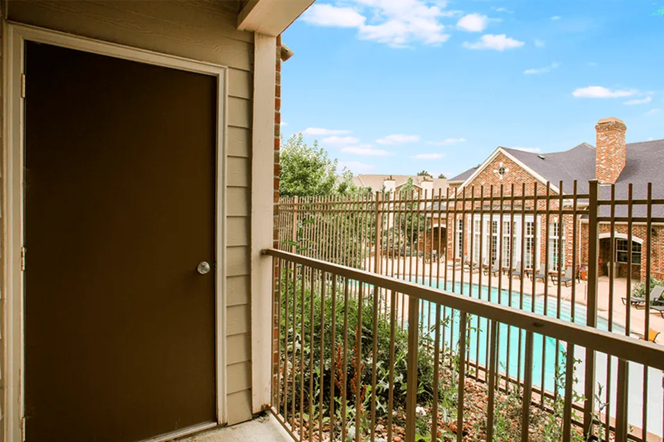 Inviting apartment balcony overlooking a luxurious pool and elegant brick clubhouse under a clear blue sky.