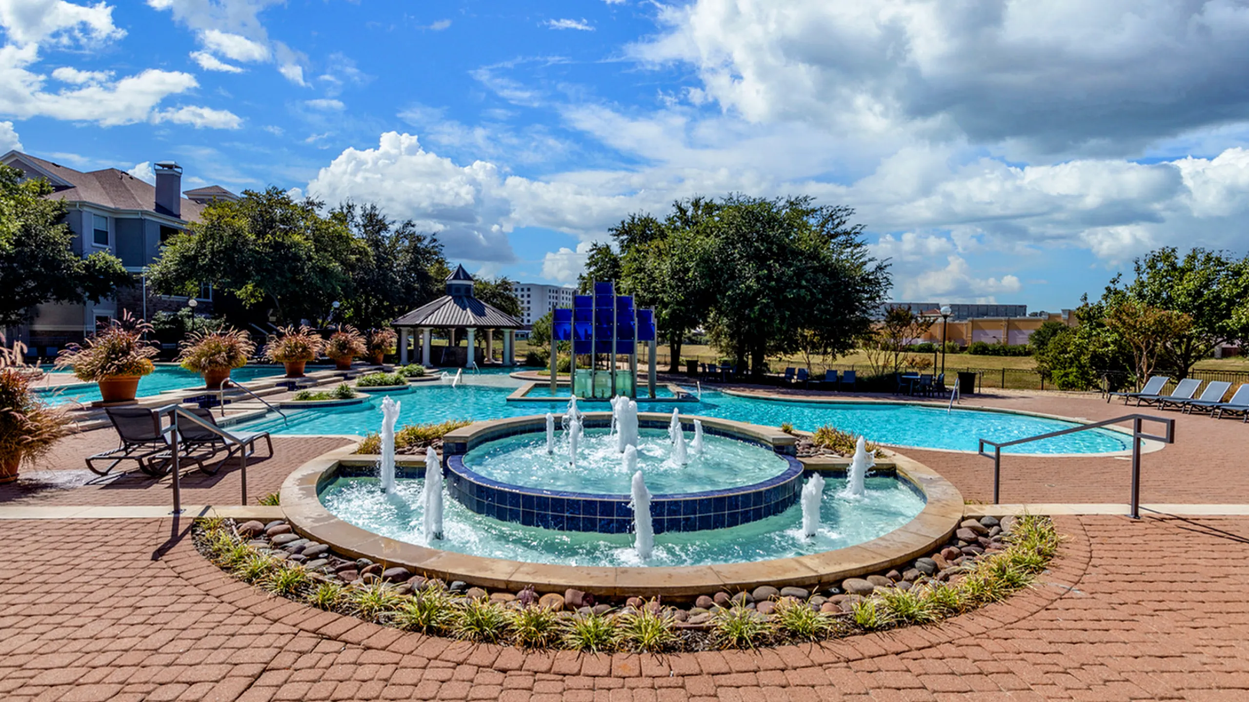 Luxurious apartment complex showcasing a vibrant outdoor pool area with elegant fountains, lush greenery, and ample sunbathing space under a bright blue sky.