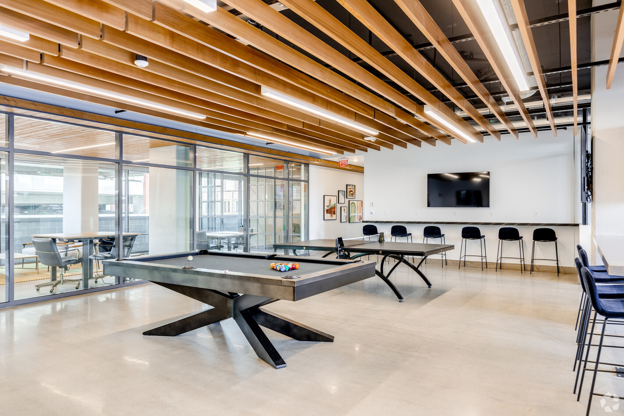 Modern recreation room with sleek billiard table, ping pong setup, and stylish seating, enhanced by wooden ceiling accents and natural light.