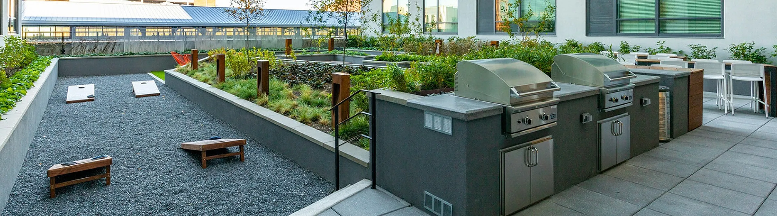 Modern apartment rooftop featuring stylish grill stations, lush greenery, and a cornhole game setup against a backdrop of city views.