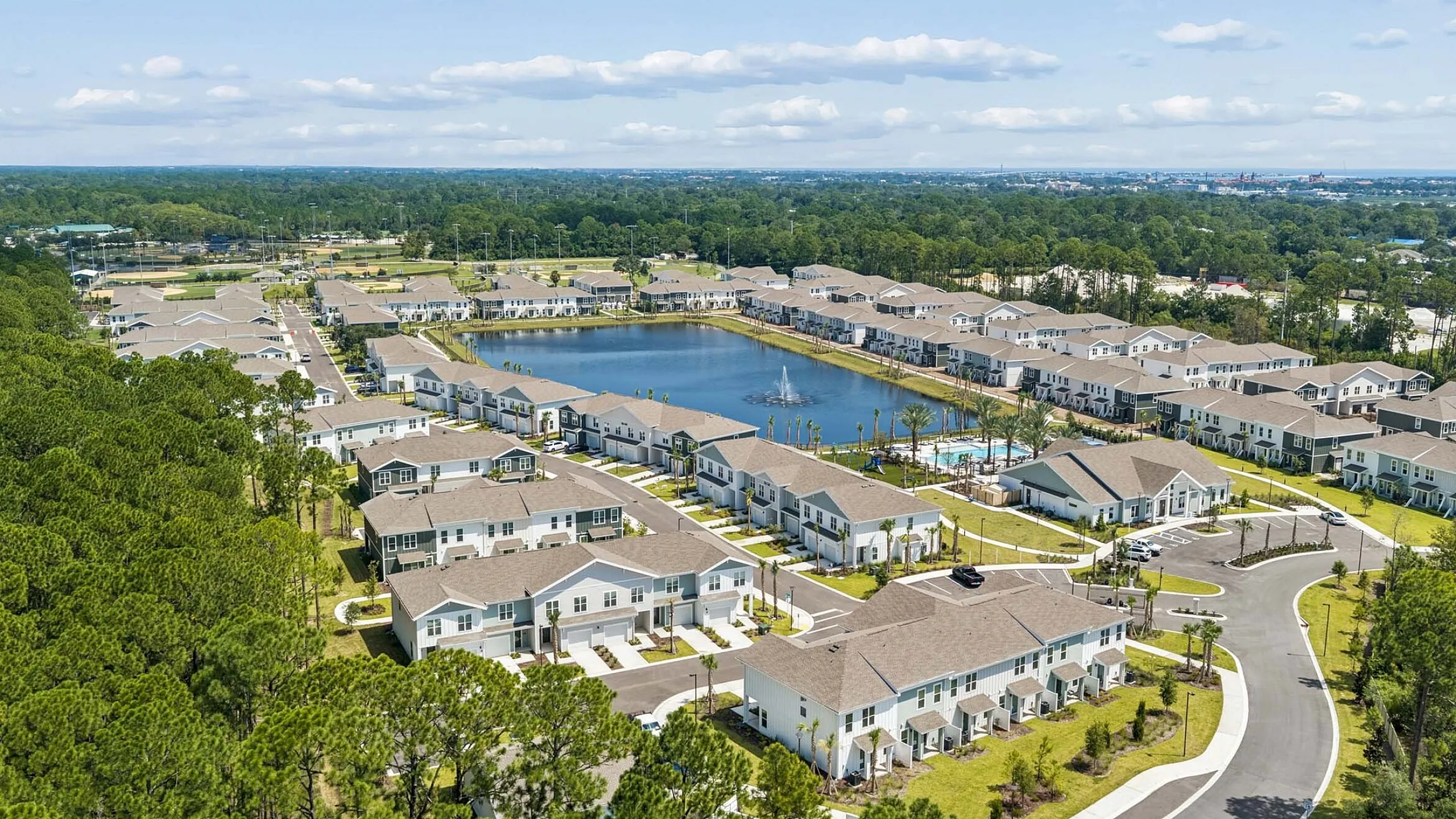 Beautiful aerial view of a modern apartment community featuring manicured landscapes, a sparkling central lake, and extensive green areas set against a lush wooded backdrop.