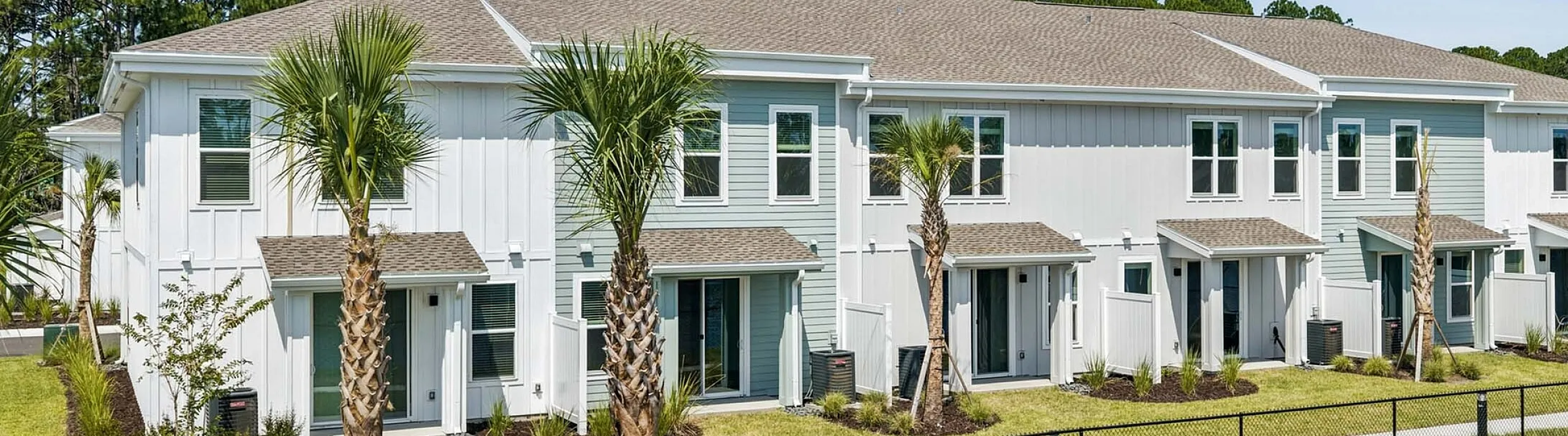 Charming modern townhomes featuring sleek white exteriors and vibrant landscaping, set against a backdrop of lush greenery and blue skies.