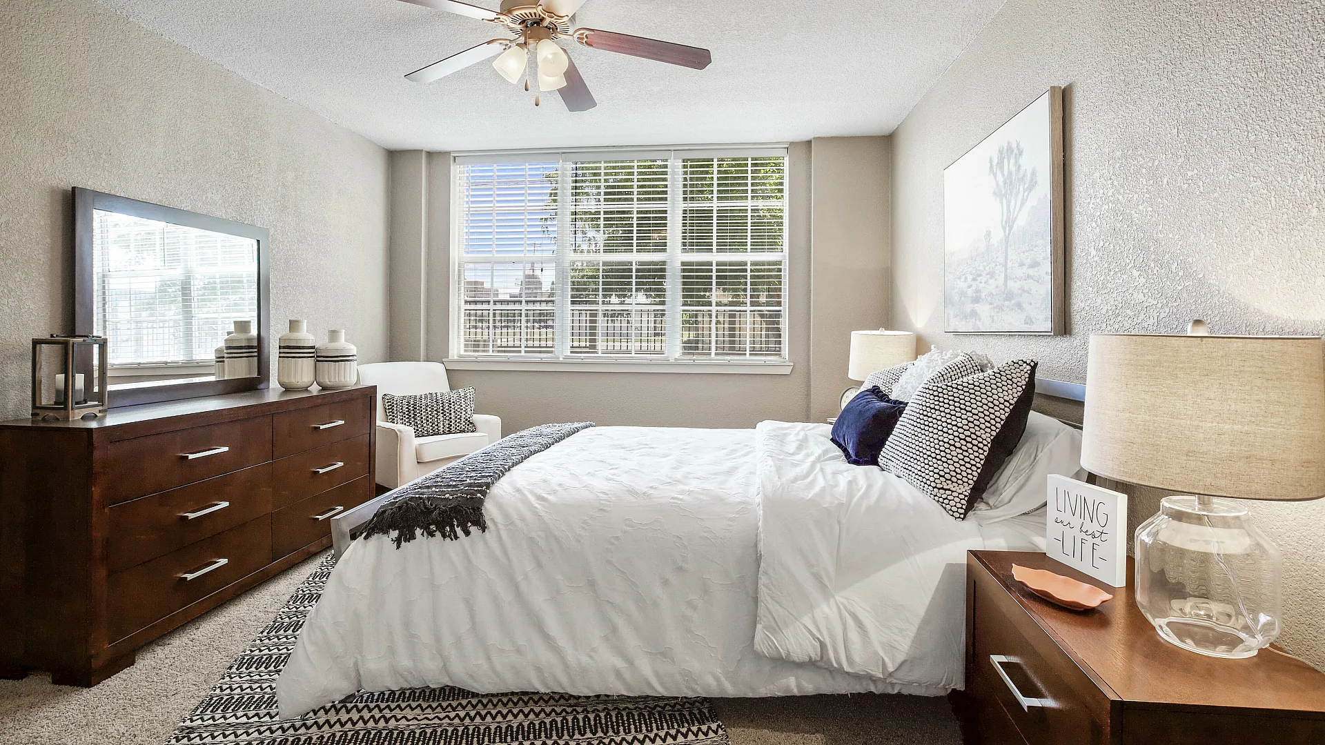 Bright and inviting bedroom with modern decor, featuring a large window, elegant ceiling fan, and cozy furnishings for ultimate comfort.